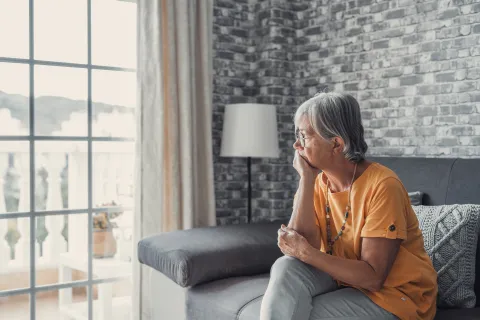 Older woman sitting on her couch and looking out the window in a thoughtful manner