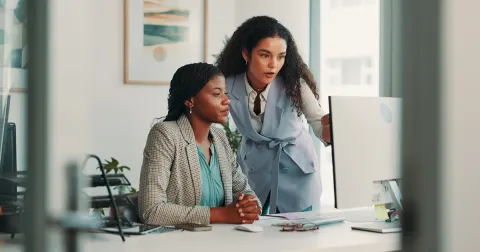 Two women in an office setting looking over and discussing what is on the computer