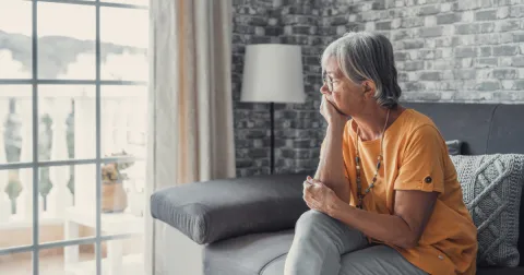 Older woman sitting on her couch and looking out the window in a thoughtful manner