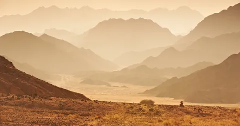 Layered mountain ranges recede into a hazy, golden desert valley at sunrise, with rocky terrain and sparse shrubs in the foreground