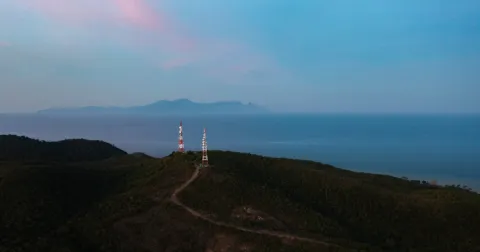 Two communication towers stand on a forested coastal ridge overlooking a calm sea, with a distant mountainous island beneath a blue and pink twilight sky